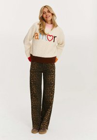 Kardigan w kolorze kremowym z wielokolorowym napisem "amor", zestawiony z brązowymi spodniami w panterkę o szerokich nogawkach i brązowymi butami.