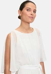 Fehér, légies ruha áttetsző köpenyujjakkal, kerek nyakkivágással és megkötő derékrésszel. Az anyag könnyűnek és texturáltnak tűnik.