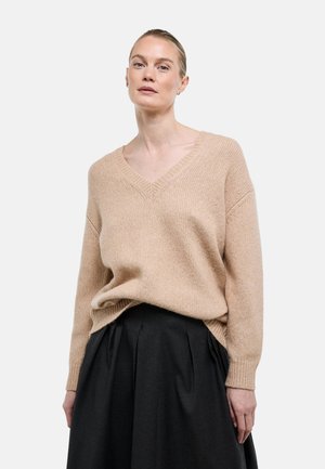 Femme aux cheveux blonds attachés porte un pull en maille beige à col en V et une jupe plissée noire devant un fond blanc uni.