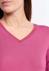 Top rosa a scollo a V realizzato in tessuto liscio, con un bordo in pizzo lavorato lungo il collo. La vista ravvicinata mette in evidenza il colore e i dettagli.