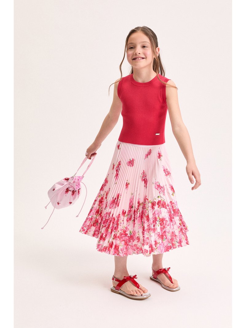 Νεαρή κοπέλα που φοράει κόκκινο τοπ χωρίς μανίκια, ροζ φλοράλ πλισέ φούστα, κόκκινες σανδάλες και κρατά μια ροζ φλοράλ τσάντα με κορδόνι.