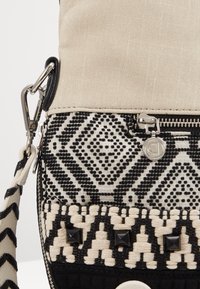 Beżowa torba z tkaniny o plecionej strukturze, z czarnymi wzorami geometrycznymi, srebrnymi elementami i teksturowanymi akcentami, z zamkiem błyskawicznym i odpinanym paskiem.