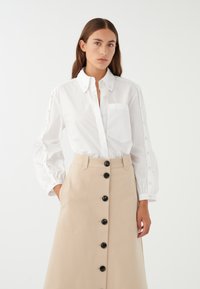 Camicia bianca con maniche lunghe a sbuffo e una piccola tasca sul petto, abbinata a una gonna A-line beige con bottoni neri sul davanti.