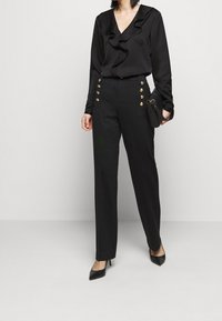 Svart blus med volangkrage och långa ärmar, i kombination med skräddarsydda svarta byxor med guldknappar på sidan. Svarta klackar fulländar looken.