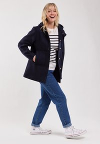 Mörkblå hooded jacka med gulddetaljer, två framfickor och en skräddarsydd passform, bärs över en vit och mörkblå randig tröja och jeans.