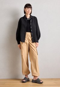 Černá košile s proužky a knoflíky, přiléhavý černý top, béžové kalhoty s elastickým pasem a zipovými kapsami a šedé boty na slip-on.