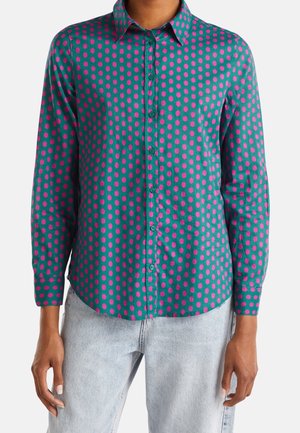 Chemise sarcelle à boutons avec des pois roses éclatants, portée avec un jean bleu clair, visible du milieu du torse jusqu’aux hanches.