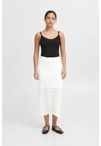 Zwarte tanktop, witte gebreide midi rok met horizontale strepen, en zwarte ronde flats; model staat tegen een neutrale achtergrond.