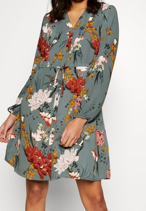 Floral dress in green with red, orange, and white flowers. Features a v-neck, button front, elasticized sleeves, and waist tie detail.