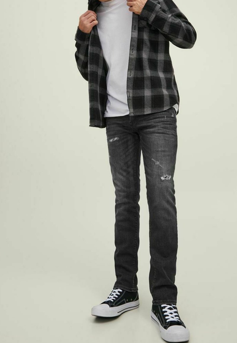 Hombre vistiendo jeans rasgados negros y grises, camisa a cuadros negra y gris sobre una camiseta blanca, y zapatillas de lona negras con cordones blancos.