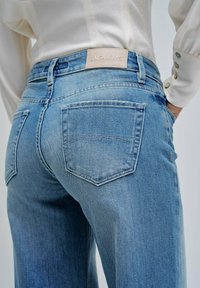 Kratak prikaz osobe koja nosi svijetloplave traperice s stražnjim džepovima i bež oznakom s natpisom "SALSA JEANS" na pojasu.