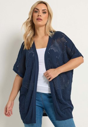 Mujer rubia vistiendo un cárdigan de punto abierto azul marino de manga corta sobre una camiseta blanca sin mangas y jeans azules, de pie frente a un fondo liso.