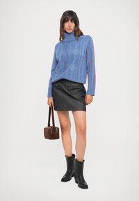 Maglione blu a collo alto lavorato a maglia a trecce con motivi a rombi, abbinato a una mini gonna di jeans nera e stivali neri alla caviglia, mentre tiene una borsa marrone.