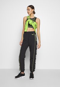 Pantaloni sportivi neri con cerniere laterali, dettagli in verde neon e vita con cordino. Abbinati a un top corto nero e a una felpa verde legata in vita.