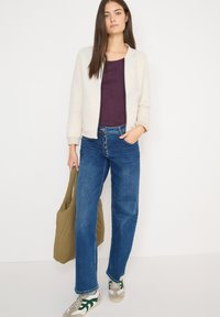 Světlá béžová bunda na zip s žebrovanou texturou, vrstvená přes tmavě fialové tričko. Modré široké džíny, šedé tenisky se zelenými doplňky a olivová taška.