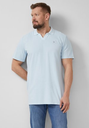 Homme avec une barbe portant un polo bleu clair et un jean bleu, debout contre un mur gris clair, regardant vers la gauche.