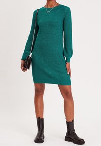 Robe verte tricotée à manches longues, texture côtelée et col en V. Associée à des bottines noires et un petit sac à main noir.