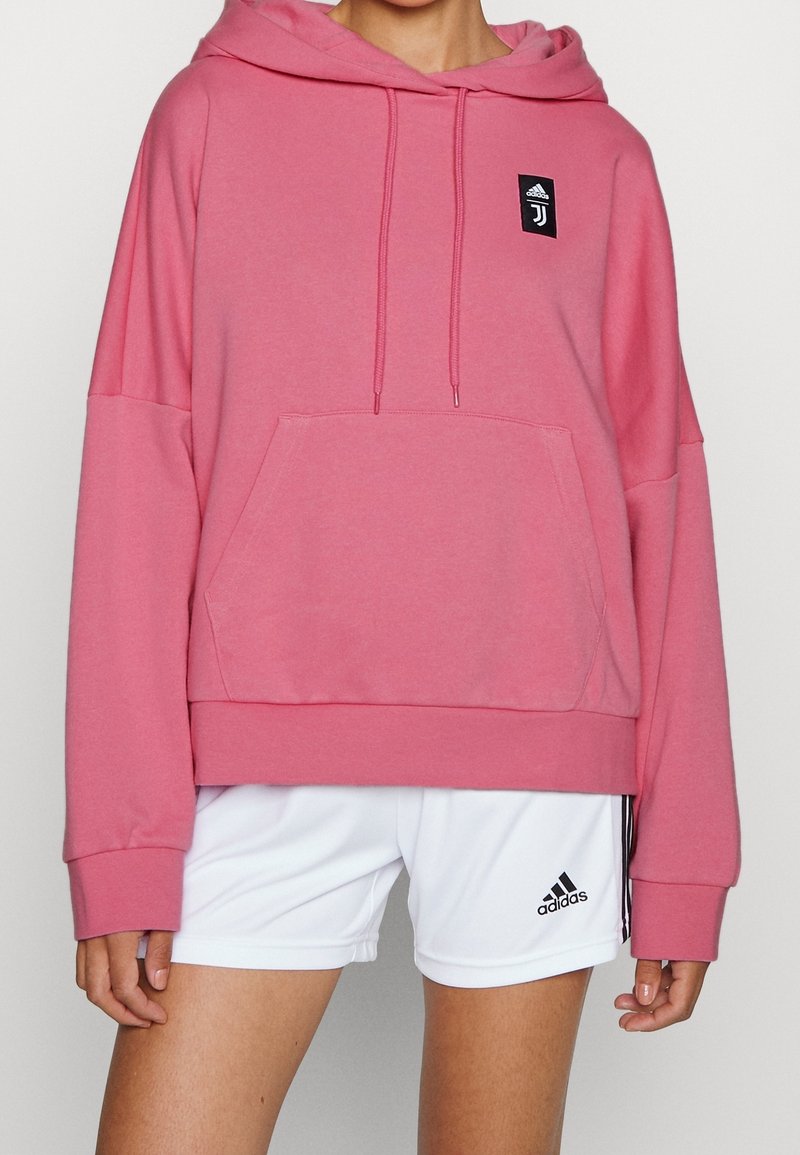 Felpa rosa con cappuccio, tasca a marsupio e polsini a costine. Presenta una patch del logo nera sul petto. Abbinata a pantaloni sporty bianchi.