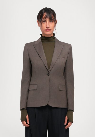 Vrouw met donker haar, gekleed in een bruine op maat gemaakte blazer over een olijfgroene top met vingerloze mouwen en donkere plissé-pantalons, staand tegen een witte achtergrond.