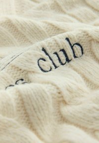 Einen cremefarbenen Strickdecken mit einem strukturierten Muster, an einer Kante mit dunkelblauem gesticktem "Club"-Text. Weiche und kuschelige Optik.