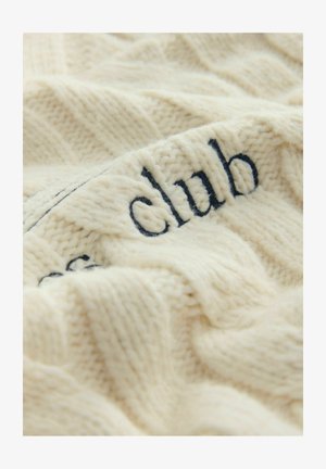 Kreemi värvi kudumite tekk, millel on tekstureeritud muster ja tumehallide "klubi"-tüüpi tikandid ühes servas. Pehme ja mugav välimus.