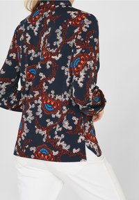 Blusa a maniche lunghe di colore blu scuro con un motivo floreale paisley in rosso, arancione e blu, caratterizzata da un orlo leggermente curvo e tessuto morbido.