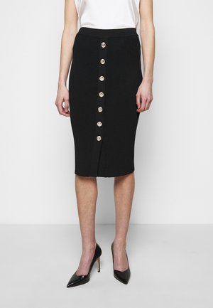 Frau trägt einen schwarzen knielangen Bleistiftrock mit silbernen Frontknöpfen und schwarze High Heels, steht vor weißem Hintergrund.