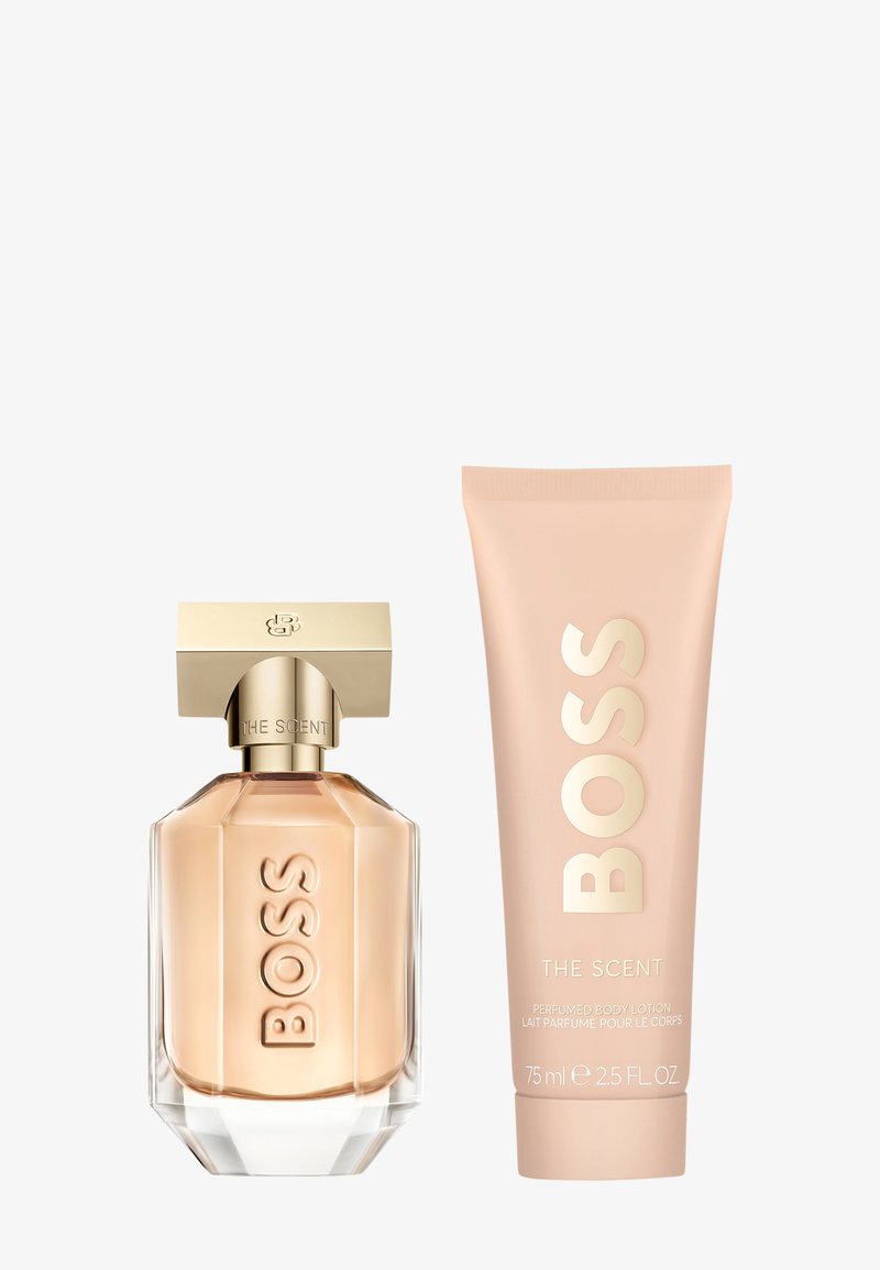 Botella de perfume de cristal con acentos dorados, base hexagonal y logotipo grabado. Junto a ella, un tubo de loción corporal rosa etiquetado "BOSS THE SCENT."