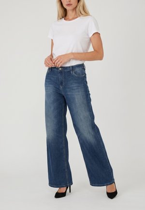 Vrouw draagt een wit T-shirt met korte mouwen, wijde blauwe jeans en zwarte hoge hakken, staand tegen een eenvoudige lichte achtergrond.