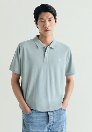 Joven con cabello negro que lleva una camiseta tipo polo azul claro y jeans azules, de pie con las manos en los bolsillos sobre un fondo liso.