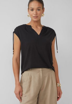 Zwart kortmouwen top met een v-hals, voorzien van plooidetails bij de schouders en koordjes met decoratieve kralen. Gecombineerd met beige broeken.