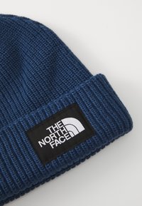 Beanie tricotat albastru cu o textură ribbed și o etichetă neagră pe care este afișat logo-ul "The North Face" în alb.