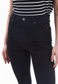 Jeans negros de cintura alta con una tela texturizada, cinco bolsillos y un cierre de botón plateado. El estilo presenta un diseño de corte ajustado.