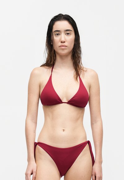 Νεαρή γυναίκα με βρεγμένα σκούρα μαλλιά, φορόντας κόκκινο μπικίνι δύο κομματιών, στέκεται μπροστά σε απλό λευκό φόντο, κοιτάζοντας προς τα εμπρός.
