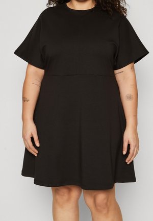 Osoba ubrana w czarną sukienkę z krótkim rękawem, pokazująca tatuaże na ramionach, stojąca na tle jednolitego, jasnego tła.