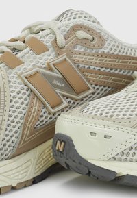 Gros plan sur des chaussures de course en mesh beige et crème avec des semelles épaisses et des logos "N" cousus bien visibles sur les côtés et les orteils.