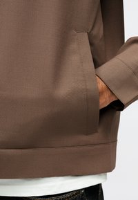 Το χέρι ενός ατόμου στην πλαϊνή τσέπη ενός καφέ σακακιού, εμφανίζοντας τις ραφές του σακακιού και μέρος ενός λευκού πουκαμίσου και σκούρου τζιν.