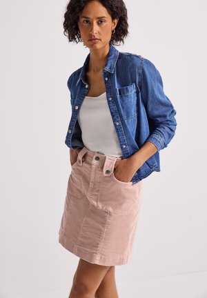 Vrouw draagt een blauwe spijkerblouse over een witte top en een lichtroze geknoopte rok met handen in de zakken, staande tegen een effen achtergrond.