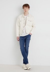 Λευκό τζάκετ από τζιν πάνω από μια λευκή μπλούζα, συνδυασμένο με μπλε τζιν και κρέμ sneakers. Το μοντέλο στέκεται μπροστά σε ανοιχτό φόντο.