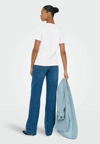 T-shirt bianco a maniche corte con scollatura rotonda, abbinato a jeans a gamba larga blu. Tiene in mano sinistra un blazer azzurro.