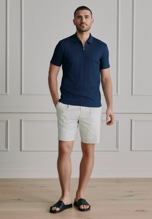 Man in navy short-sleeve polo shirt, white shorts, and black slide sandals standing with one hand in pocket against white paneled wall.