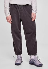 Pantaloni grigio scuro a vestibilità comoda con vita e polsini elastici; tessuto liscio; indossati con scarpe da corsa viola chiaro con design a rete.