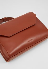 Borsa a mano in pelle marrone con chiusura a patta, texture liscia, forma rettangolare e branding goffrato discreto sul davanti.