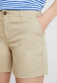 Beige bomuldsshorts med fladt foran, sidelommer og knaplukning. Stoffet har en glat tekstur og en ren finish.