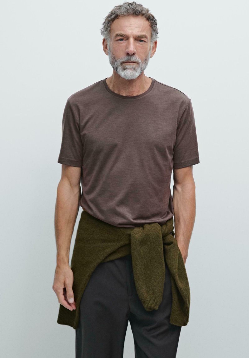 Brun kortærmet T-shirt med rund halsudskæring, parret med mørke bukser og en grøn sweater bundet om taljen, stående mod en neutral baggrund.