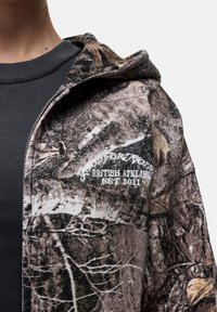 Felpa con zip in camouflage con un tessuto testurizzato; presenta un cappuccio, uno strato interno scuro e un logo ricamato bianco sul petto.