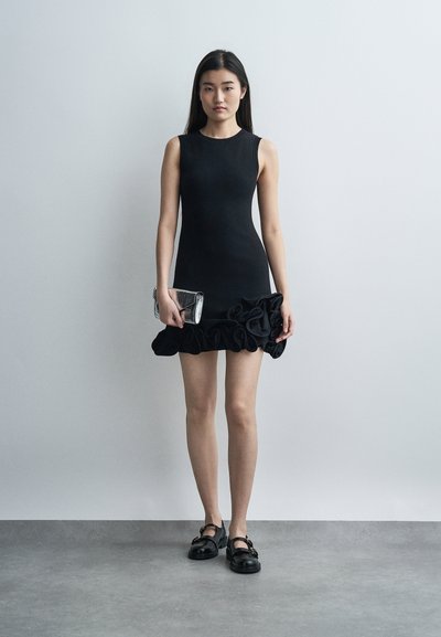 Γυναίκα που φοράει ένα μαύρο μίνι φόρεμα χωρίς μανίκια με βολάν στο τελείωμα, κρατώντας μια ασημένια τσάντα χειρός και μαύρα παπούτσια, στέκεται μπροστά σε γκρίζο τοίχο.