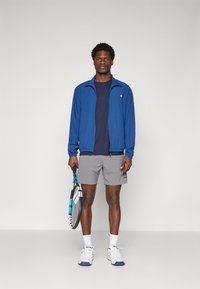 Kék cipzáras sportdzseki, tengerészkék póló, szürke rövidnadrág, fehér zokni és teniszcipő. Kék akcentusokkal ellátott teniszütőt tart a kezeiben.