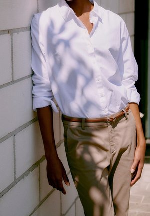 Osoba opierająca się o jasno szarą ceglany mur, ubrana w białą koszulę zapinaną na guziki i beżowe spodnie z brązowym paskiem, cienie padające na ubranie.