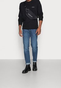 Borsa a tracolla nera con logo, indossata sopra un maglione scuro, abbinata a jeans azzurri chiari e stivali neri alla caviglia, con i risvolti.